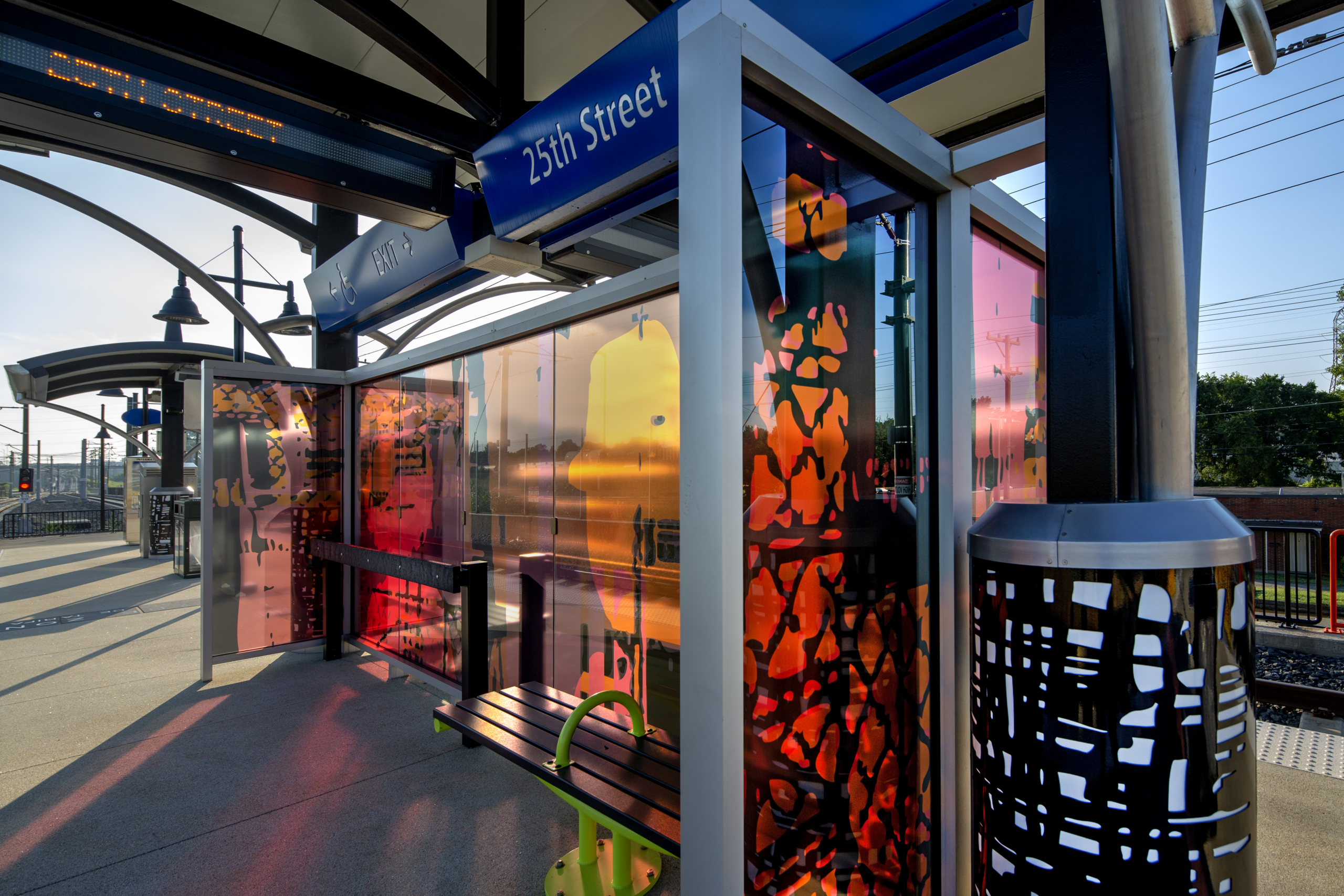 Glass windscreen with laminated black, pink, & orange patterns & human silhouettes as through a passing train window cast colorful shadows on the 25th St Station light rail platform. Canopy columns with b&w metal abstract designs complement the windscreen.