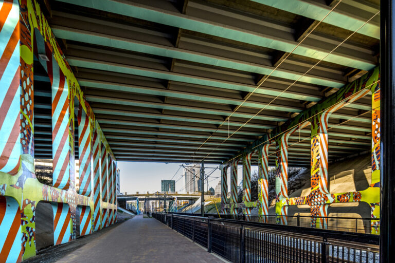 Detail of I-277 & 11th St underpass over light rail track & walkway in Charlotte. Sidewalk perspective looking down track towards city. Mural painted sky blue with cobalt blue & orange diagonal, repeating, lined patterns down sides of concrete support ballasts. Mesh-like & organic patterns in various bright colors throughout.