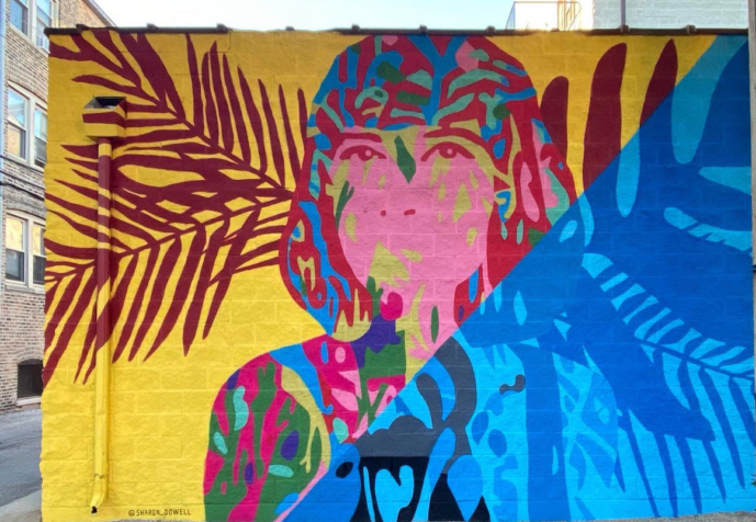 15” h x 24’ w mural on exterior concrete wall of woman painted in vibrant colors with shoulder-length hair facing forward, looking outward. Mural bisected diagonally: yellow background with maroon palm fronds on left side, right side blocked in all blues. In shadows of Ashland Ave L Train bridge, Lakeview Chicago.