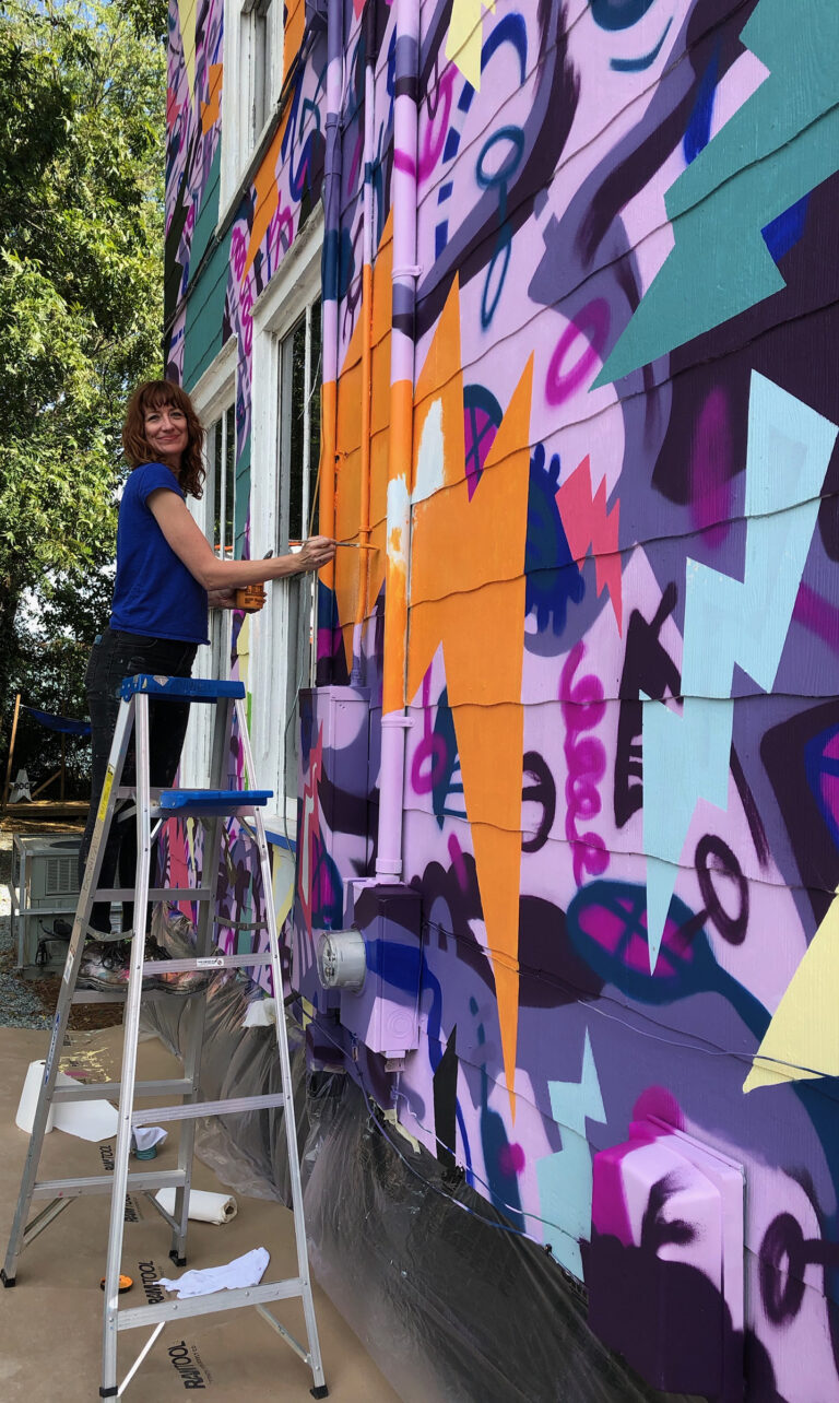 Sharon Dowell painting a colorful, two-story mural of comic book-style lightning bolts & playful shapes & lines covering the entire exterior wall of Girls Rock CLT headquarters.