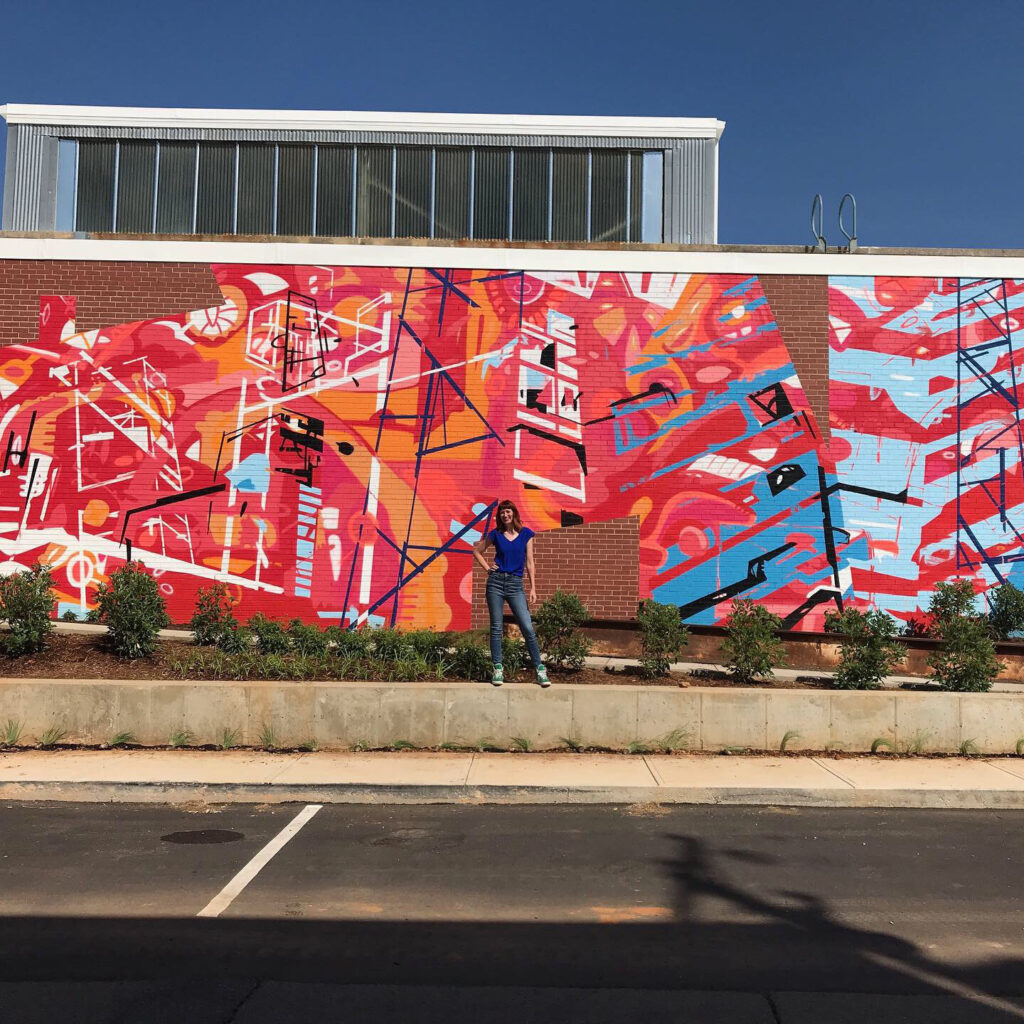 20’ h x 80’ w hand & spray-painted energetic, abstract mural with overlapping planes of reds, blues & oranges. Architectural lines in blues, black & white echo industrial buildings and towers of surroundings. Precise geometric shapes of exposed, unpainted red brick. Located on exterior wall of Goodyear Arts building in Charlotte’s Camp North End.