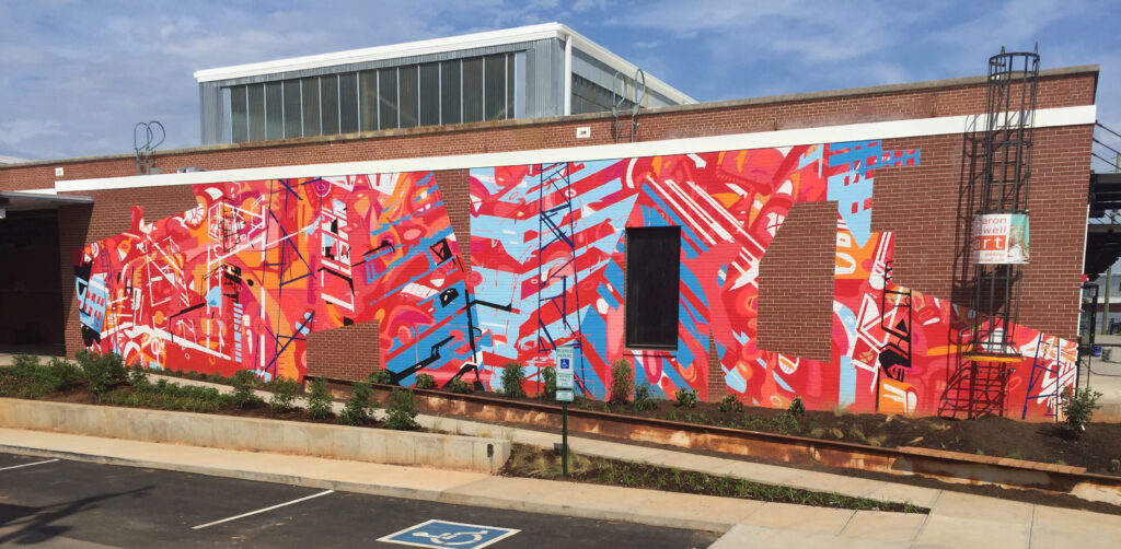 20’ h x 80’ w hand & spray-painted energetic, abstract mural with overlapping planes of reds, blues & oranges. Architectural lines in blues, black & white echo industrial buildings and towers of surroundings. Precise geometric shapes of exposed, unpainted red brick. Located on exterior wall of Goodyear Arts building in Charlotte’s Camp North End.