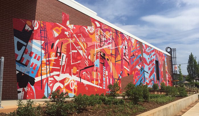 20’ h x 80’ w hand & spray-painted energetic, abstract mural with overlapping planes of reds, blues & oranges. Architectural lines in blues, black & white echo industrial buildings and towers of surroundings. Precise geometric shapes of exposed, unpainted red brick. Located on exterior wall of Goodyear Arts building in Charlotte’s Camp North End.