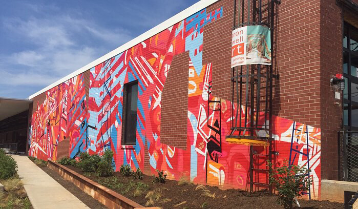 20’ h x 80’ w hand & spray-painted energetic, abstract mural with overlapping planes of reds, blues & oranges. Architectural lines in blues, black & white echo industrial buildings and towers of surroundings. Precise geometric shapes of exposed, unpainted red brick. Located on exterior wall of Goodyear Arts building in Charlotte’s Camp North End.