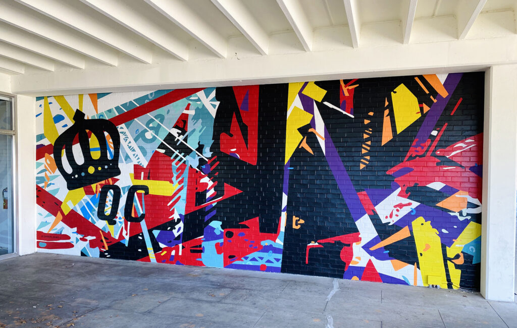 Energetic mural of geometric shapes & abstracted trusses & beams in bold colors on brick wall. Charlotte NC’s crown logo and QC painted prominently in black on left side of mural. Located outside Bart’s Bottle Shop, Eastway Shopping Center.