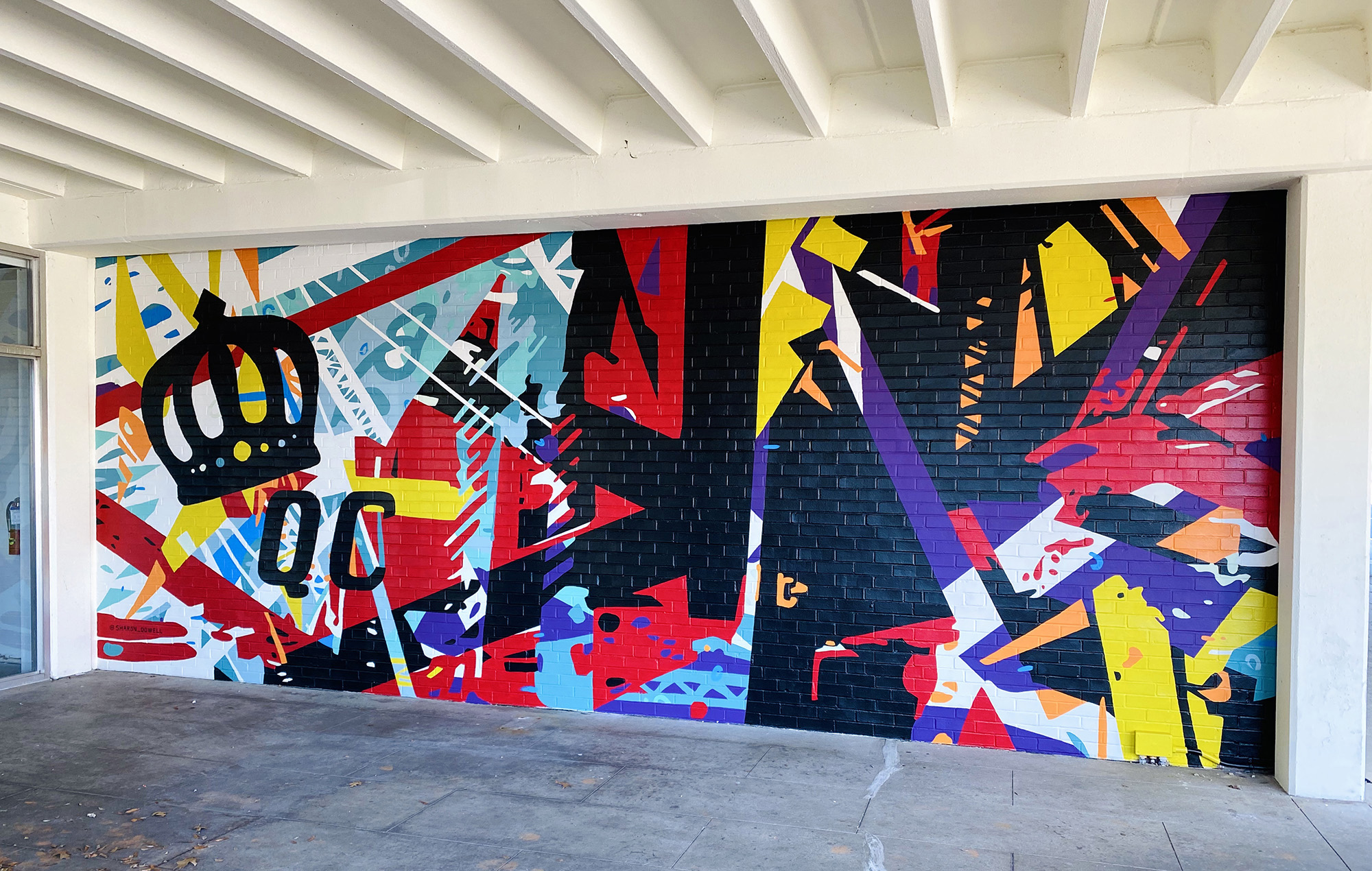 Energetic mural of geometric shapes & abstracted trusses & beams in bold colors on brick wall. Charlotte NC’s crown logo and QC painted prominently in black on left side of mural. Located outside Bart’s Bottle Shop, Eastway Shopping Center.