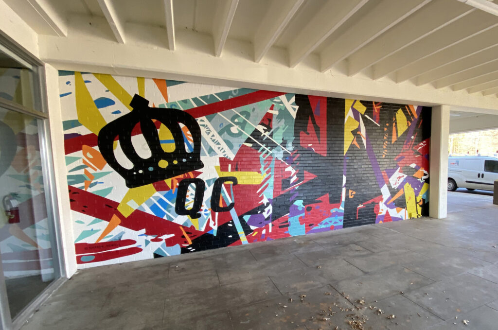 Energetic mural of geometric shapes & abstracted trusses & beams in bold colors on brick wall. Charlotte NC’s crown logo and QC painted prominently in black on left side of mural. Located outside Bart’s Bottle Shop, Eastway Shopping Center.