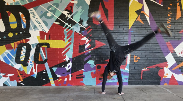 Energetic mural of geometric shapes & abstracted trusses & beams in bold colors on brick wall. Charlotte NC’s crown logo and QC painted prominently in black on left side of mural. Located outside Bart’s Bottle Shop, Eastway Shopping Center.