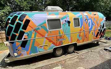 Aluminum Airstream Argosy completely painted in enamel, with images of tools on a brightly-colored background of geometric shapes. Large She Built This City logo in black on side. United We Spark in teal block letters over rear windshield.