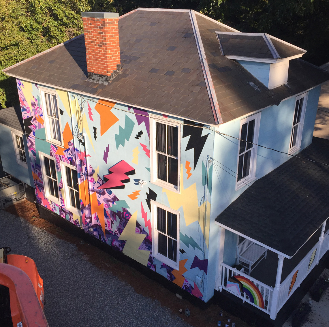 A colorful, two-story, hand-painted mural of comic book-style lightning bolts & playful shapes & lines covering the entire exterior wall of Girls Rock CLT headquarters. Summer camp volunteers and youth also helped create the inclusivity rainbow and painted, aluminum lighting bolt sculptures on front porch railing.