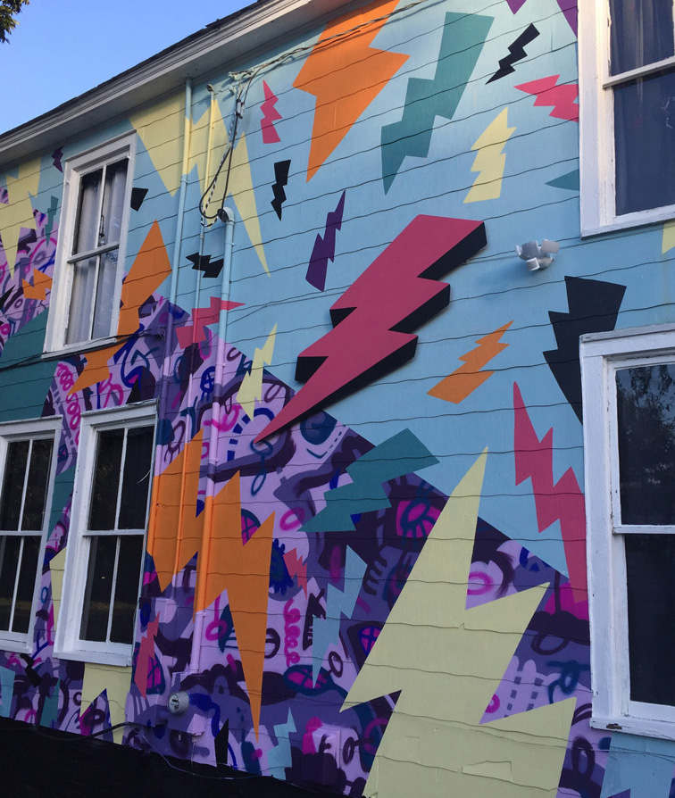 A colorful, two-story, hand-painted mural of comic book-style lightning bolts & playful shapes & lines covering the entire exterior wall of Girls Rock CLT headquarters. Summer camp volunteers and youth also helped create the inclusivity rainbow and painted, aluminum lighting bolt sculptures on front porch railing.