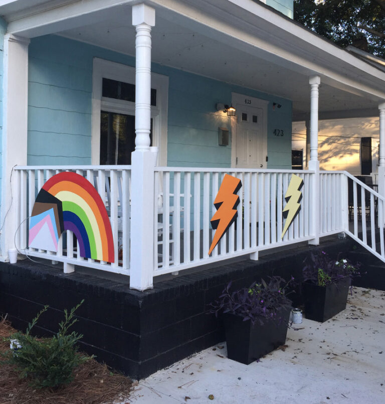 A colorful, two-story, hand-painted mural of comic book-style lightning bolts & playful shapes & lines covering the entire exterior wall of Girls Rock CLT headquarters. Summer camp volunteers and youth also helped create the inclusivity rainbow and painted, aluminum lighting bolt sculptures on front porch railing.