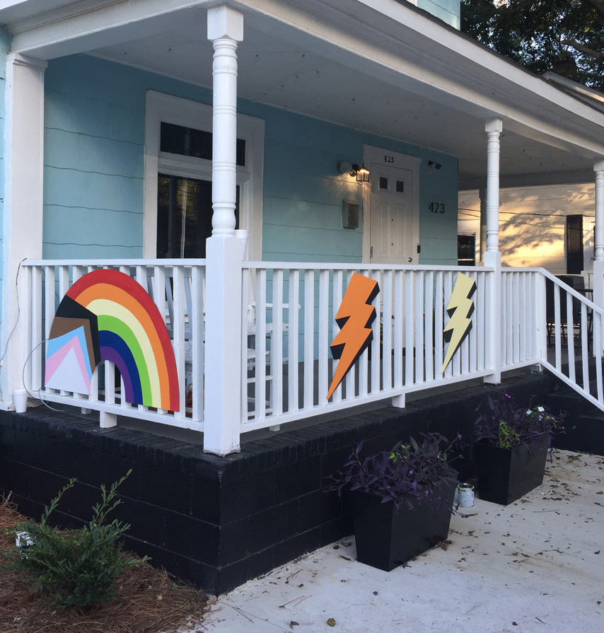 A colorful, two-story, hand-painted mural of comic book-style lightning bolts & playful shapes & lines covering the entire exterior wall of Girls Rock CLT headquarters. Summer camp volunteers and youth also helped create the inclusivity rainbow and painted, aluminum lighting bolt sculptures on front porch railing.