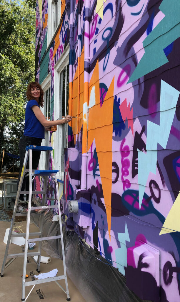 A colorful, two-story, hand-painted mural of comic book-style lightning bolts & playful shapes & lines covering the entire exterior wall of Girls Rock CLT headquarters. Summer camp volunteers and youth also helped create the inclusivity rainbow and painted, aluminum lighting bolt sculptures on front porch railing.