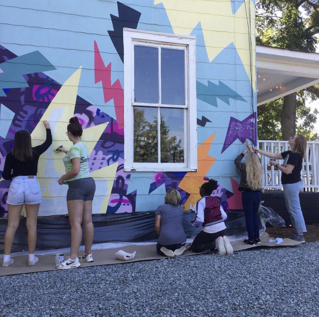 A colorful, two-story, hand-painted mural of comic book-style lightning bolts & playful shapes & lines covering the entire exterior wall of Girls Rock CLT headquarters. Summer camp volunteers and youth also helped create the inclusivity rainbow and painted, aluminum lighting bolt sculptures on front porch railing.