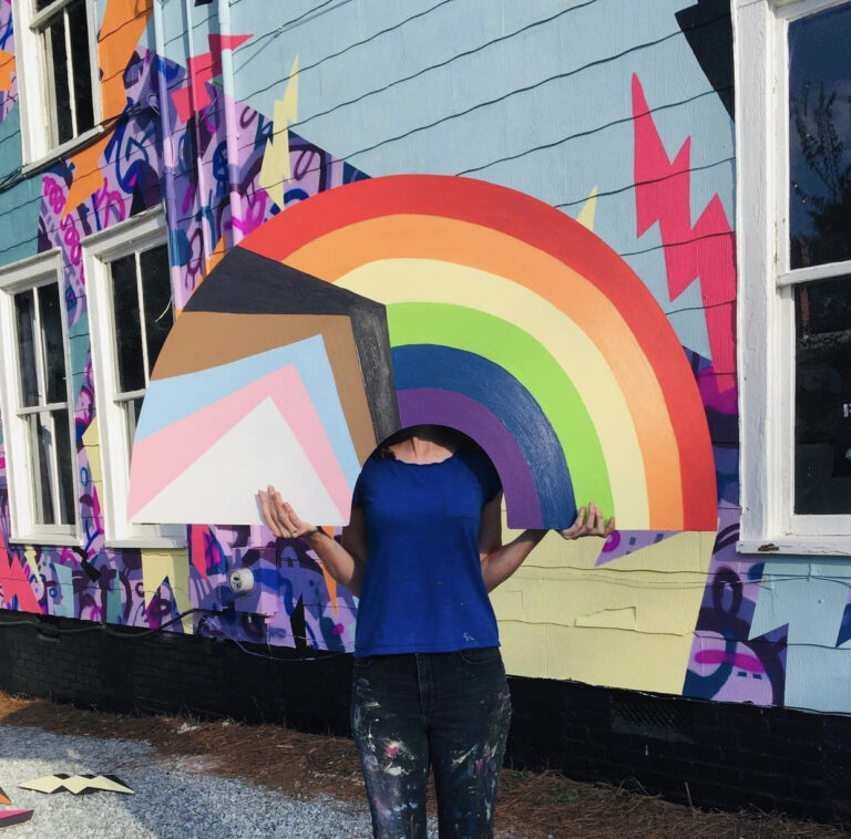 A colorful, two-story, hand-painted mural of comic book-style lightning bolts & playful shapes & lines covering the entire exterior wall of Girls Rock CLT headquarters. Summer camp volunteers and youth also helped create the inclusivity rainbow and painted, aluminum lighting bolt sculptures on front porch railing.